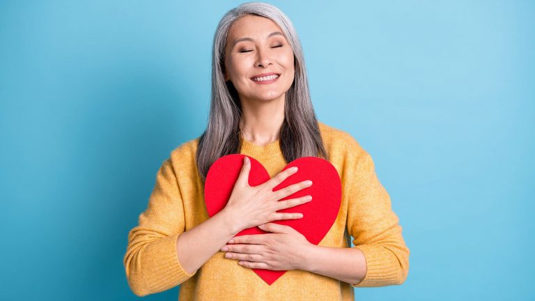 Photograph Of Grey-Haired Woman Smiling While Hugging Giant Red Heart Cut From Cardboard