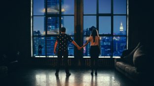 Photograph Of Man And Woman Standing Apart While Holding Hands And Looking Over A Cityscape From Their Apartment