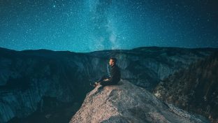 Photograph Of Lone Man Sitting On Rock Cliff Beneath Starry Sky With Milky Way Visible