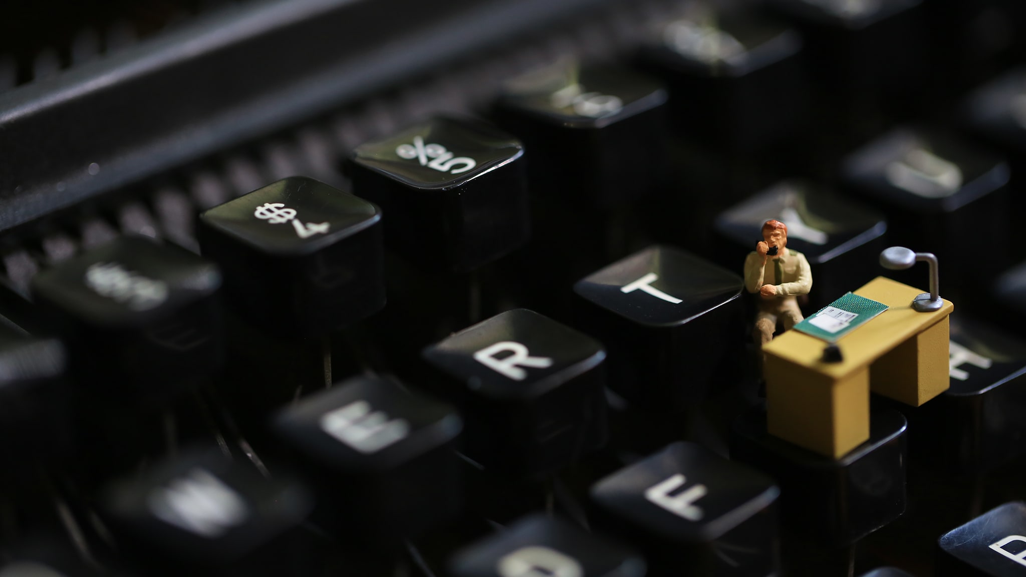 Zoomed In View Of A Black Keyboard With A Tiny Desk And Person Sitting At The Desk On One Of The Keys Signifying Workplace Loneliness And Isolation