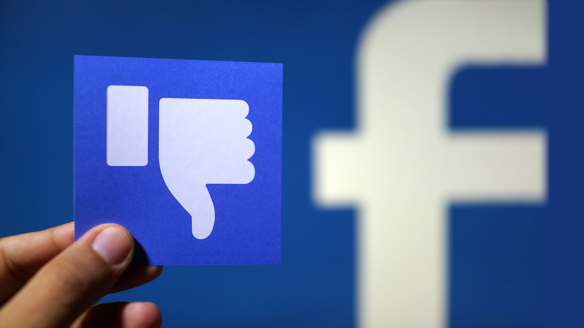 Photograph Of Hand Holding Small Card With "Thumbs Down" Sign With Part Of Facebook's Logo Blurred In The Background