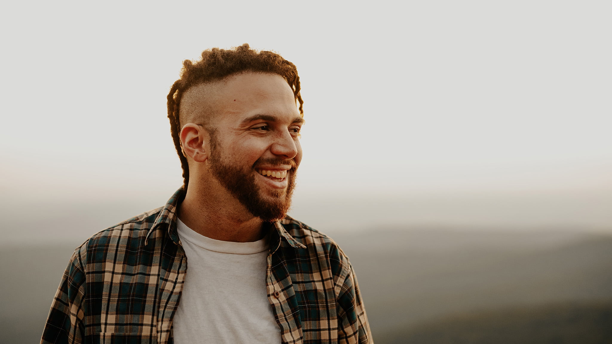 Photograph Of Middle-Aged Man Wearing A Flannel Shirt And Looking Off-Camera, Smiling With Misty Mountain Background