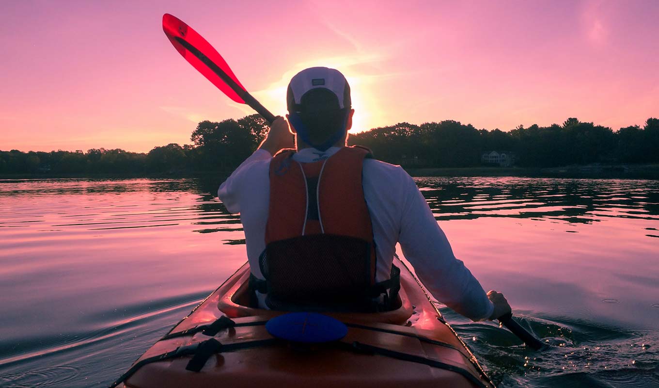 Rear View Silhouette Of Man Paddling Kayak Across Sunset Lake