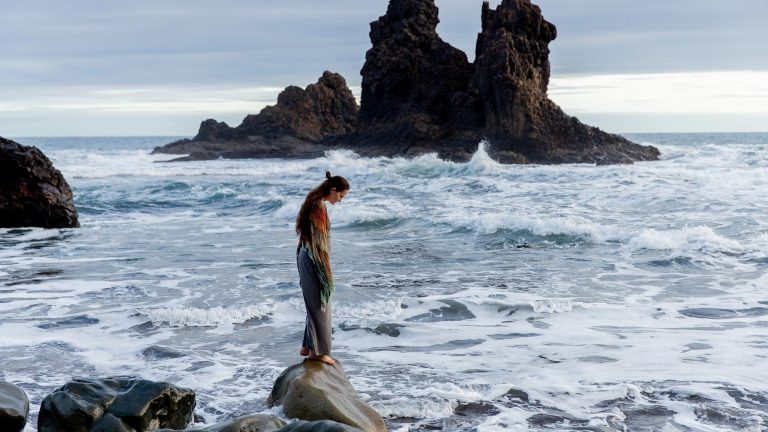 melancholy photograph of a woman standing alone on a rock along the shoreline, looking down at the tumultuous waves surrounding her