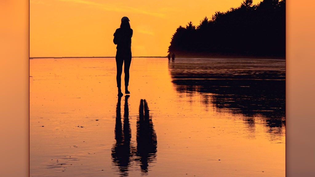 moody sunset photograph of woman walking along the beach with her shadow behind her, accompanied by only her husband's shadow on the ground, widow standing alone in grief concept