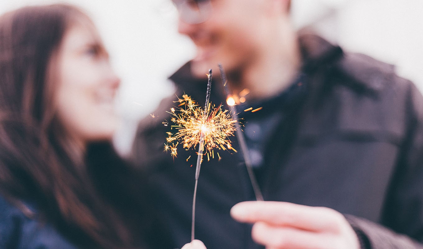 Photograph Of Two Lit Sparklers With A Smiling Couple In The Out-Of-Focus Background