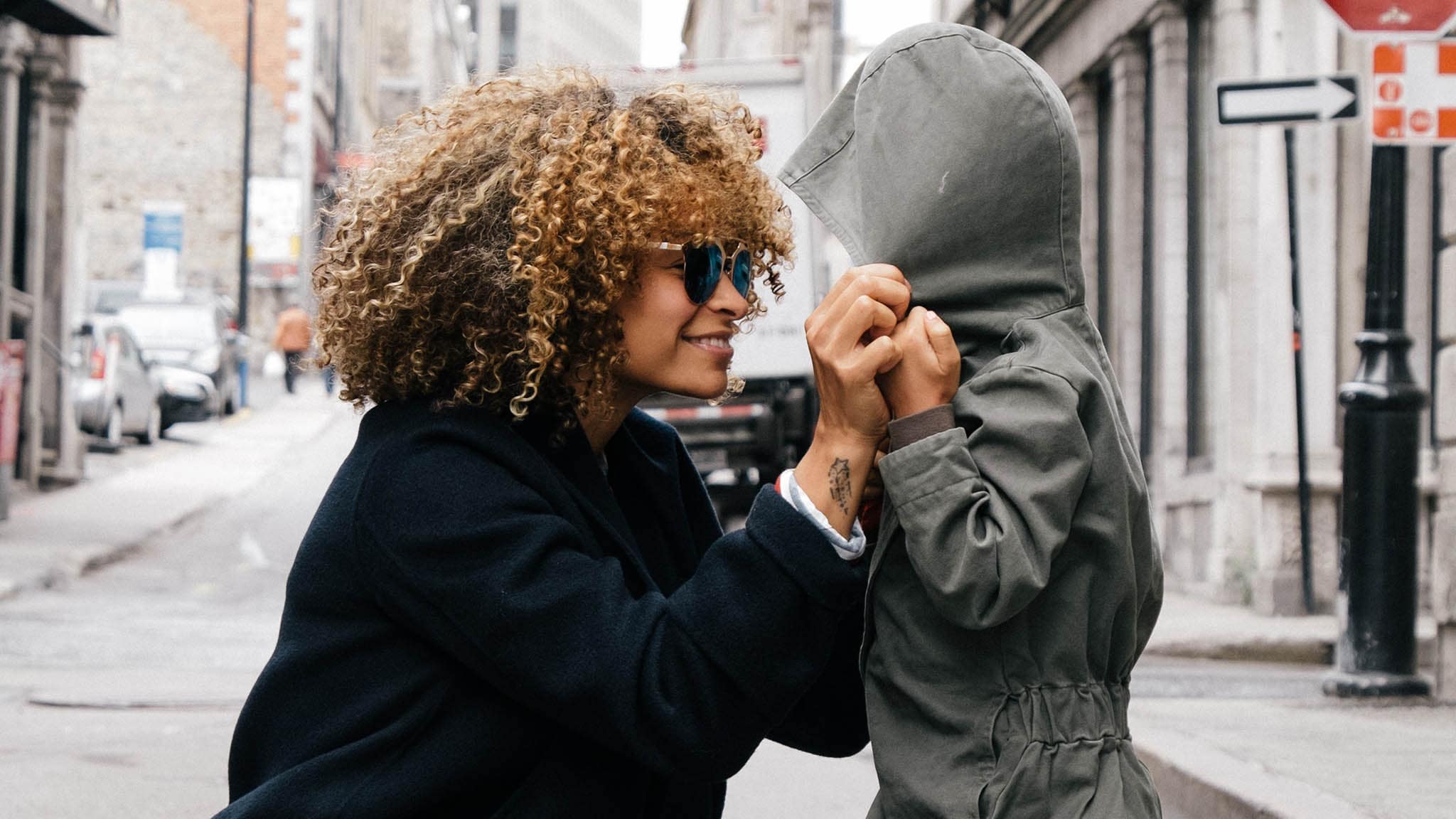 Why Parenting Can Be Lonely, Even When You're Rarely Alone Side View Outdoor Photograph Of Woman Smiling While Kneeling And Straightening A Young Child's Hood