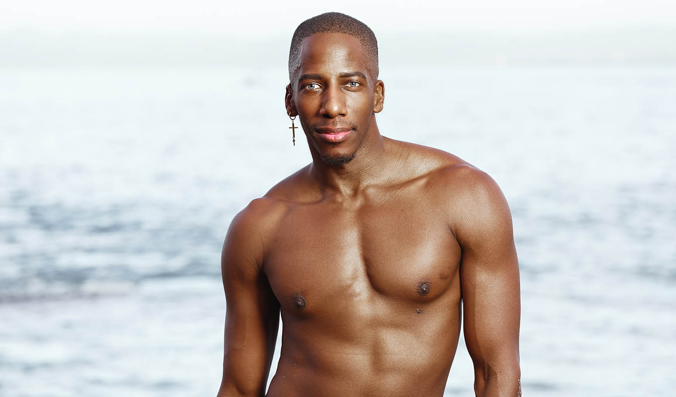 Photograph Of Dark-Skinned Male Model Standing Shirtless In Front Of A Body Of Water