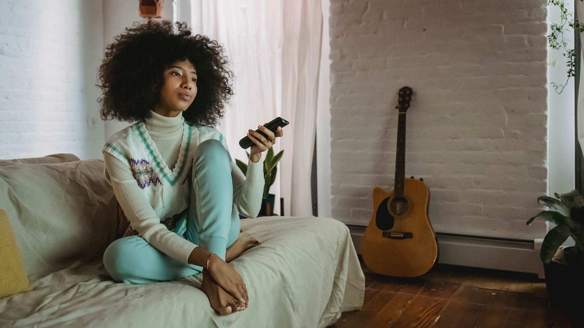 Photograph Of Young Woman Sitting Alone On Couch While Absentmindedly Hitting Buttons On Remote Control