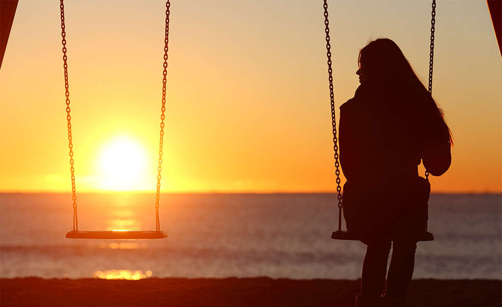 photograph featuring the silhouette of a woman sitting on a swing set alone at sunset while looking toward the empty swing beside her to convey grief and loss