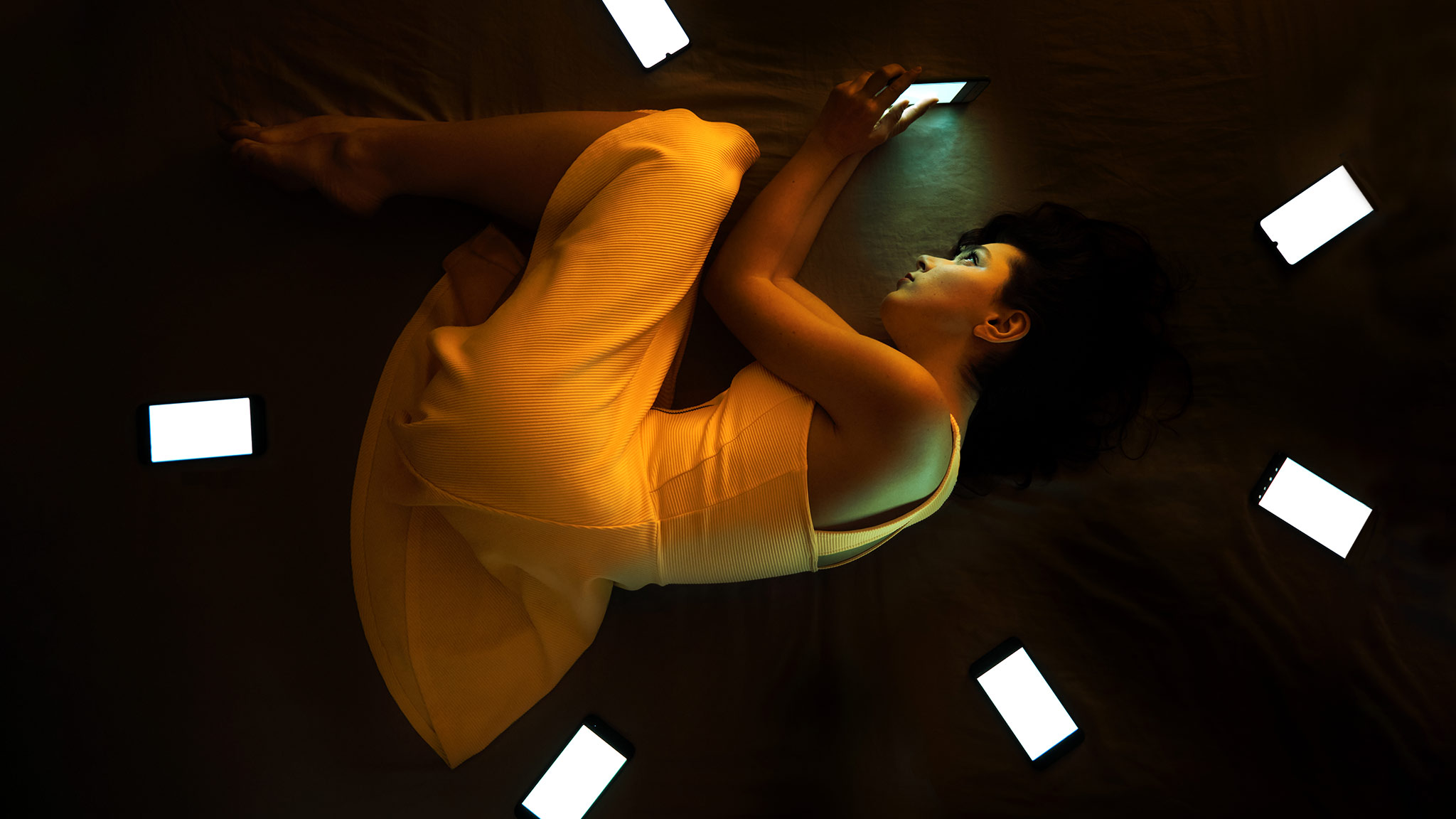 Aerial Photograph Of Woman Laying On Bed Scrolling Through Her Phone, Surrounded By Other Lit Phone Screens In Every Direction