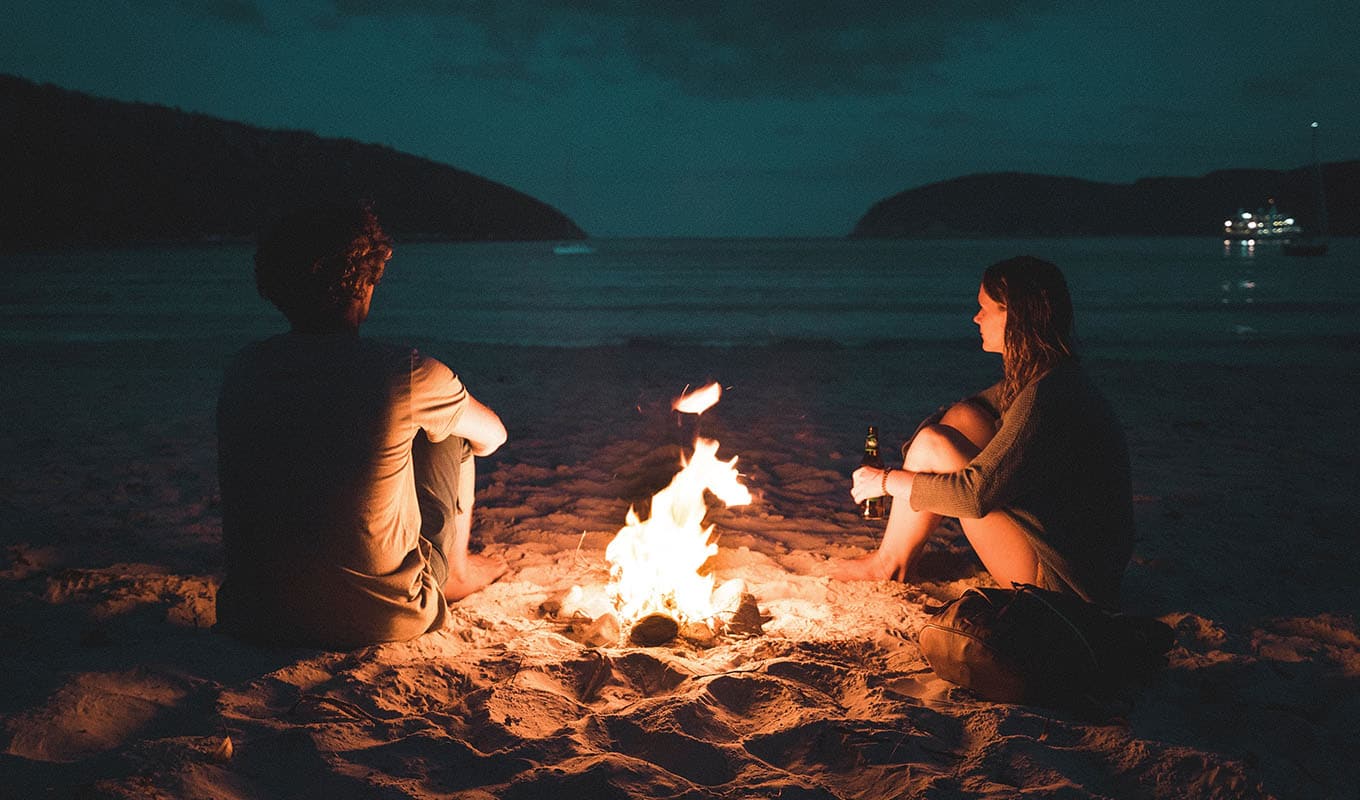 Photograph Of Man And Woman Sitting In The Sand Around A Campfire And Looking Out Over The Water
