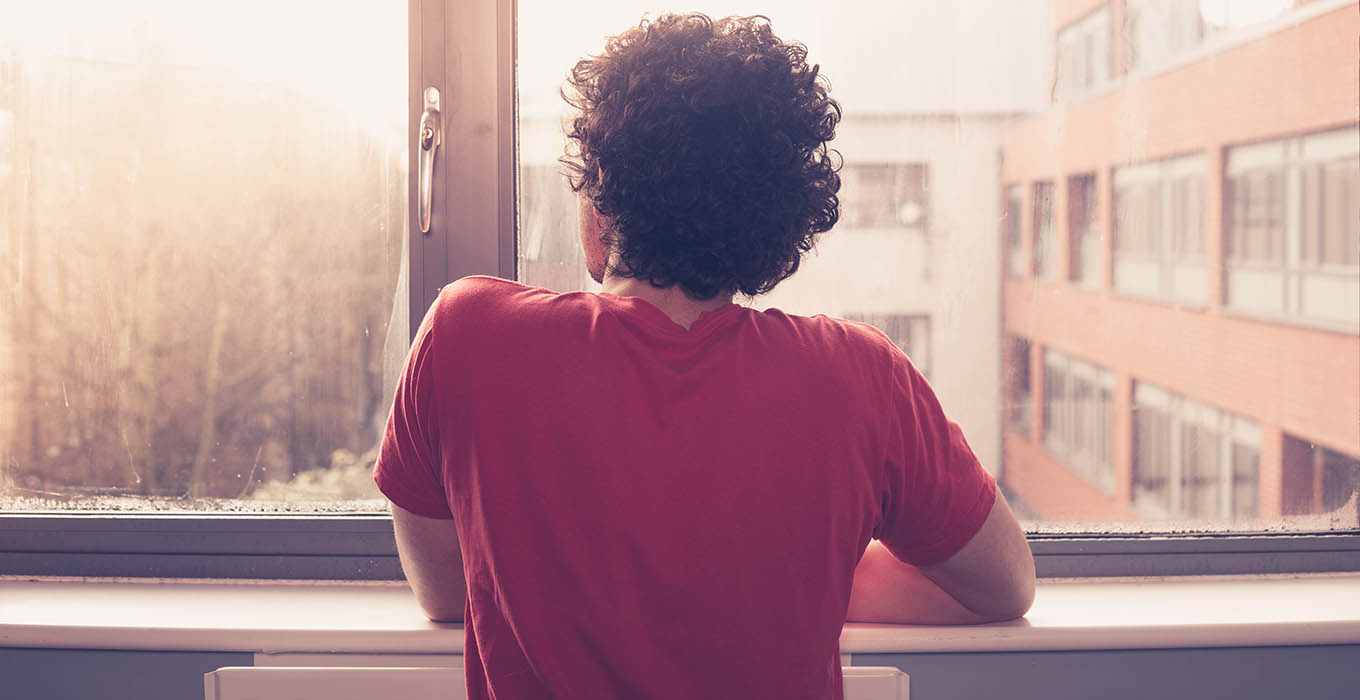 photo of young man rear view looking out the window lamenting quarantine loneliness