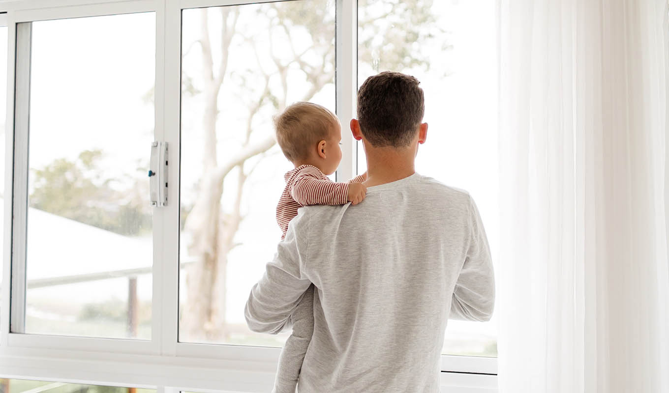Rear View Indoor Photograph Of Man Holding Baby As They Look Outside Through A Picture Window