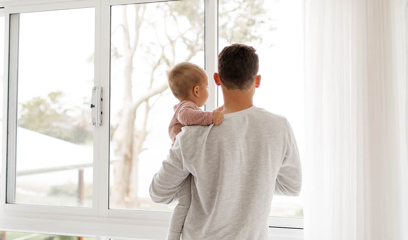 photograph of a male parent holding their baby and looking out the window, experiencing loneliness in the journey of parenthood