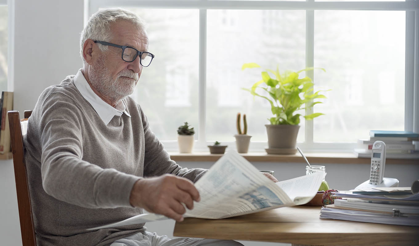 photo of a senior man reading a newspaper in the kitchen while living alone