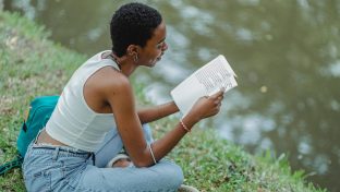 photograph of woman sitting in the grass along the water's edge, reading a handwritten journal